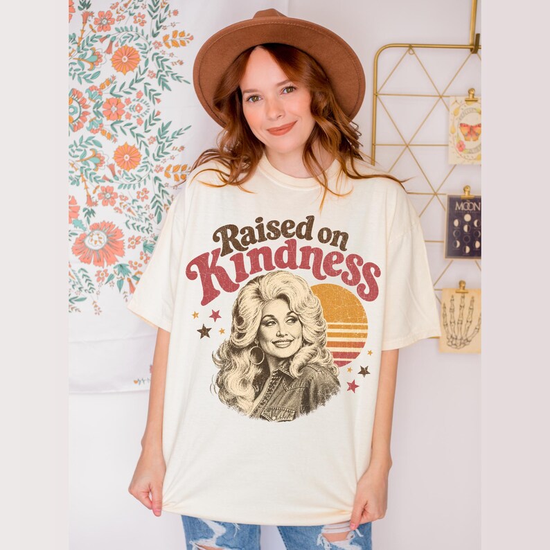 May include: A cream-colored short-sleeve t-shirt featuring a graphic print of a woman with voluminous blonde hair. The text "Raised on Kindness" is arched above the portrait, accompanied by a sunset graphic and small stars in red and brown tones.