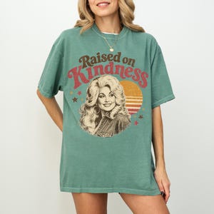 May include: A sage green short-sleeve t-shirt featuring a graphic print of a woman with voluminous blonde hair. The text "Raised on Kindness" is printed in red above the portrait, which is set against a circular orange sun with horizontal lines.