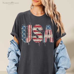 May include: Dark gray t-shirt with the word "USA" in a distressed, patriotic design. The letters feature the American flag's red and white stripes and blue stars. The shirt has rolled-up sleeves and is part of a casual clothing collection.