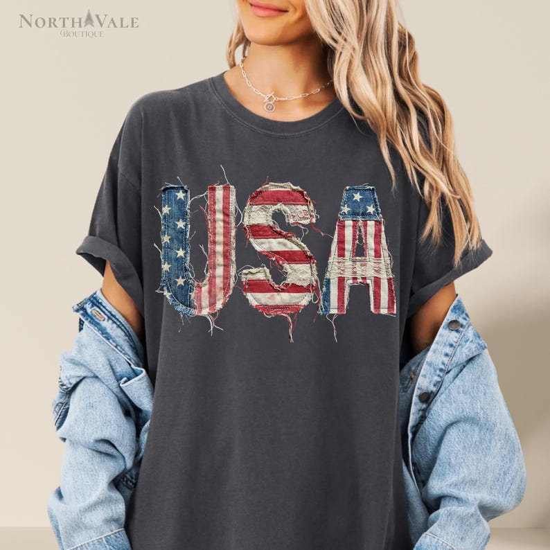 May include: Dark gray t-shirt with the word "USA" in a distressed, patriotic design. The letters feature the American flag's red and white stripes and blue stars. The shirt has rolled-up sleeves and is part of a casual clothing collection.