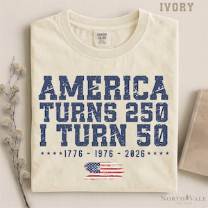 May include: Ivory-colored t-shirt with the text "AMERICA TURNS 250 I TURN 50" in distressed blue lettering. Includes the years 1776, 1976, and 2026, along with an American flag graphic. A Comfort Colors brand shirt.