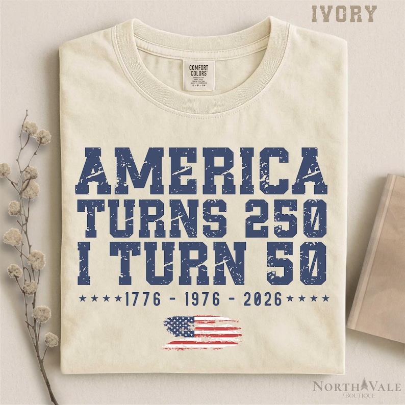May include: Ivory-colored t-shirt with the text "AMERICA TURNS 250 I TURN 50" in distressed blue lettering. Includes the years 1776, 1976, and 2026, along with an American flag graphic. A Comfort Colors brand shirt.