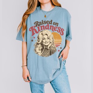 May include: A dusty blue short-sleeve t-shirt featuring a graphic print of a woman with voluminous blonde hair. The text "Raised on Kindness" is arched above the portrait, accompanied by a sunset circle and small star icons in red and gold.