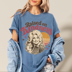 May include: A blue short-sleeve t-shirt featuring a graphic print of a woman with voluminous blonde hair. The text "Raised on Kindness" is printed in brown and red above the portrait, accompanied by a yellow sun graphic and small stars.