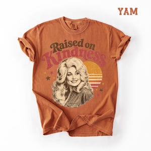 May include: A burnt orange short-sleeve t-shirt featuring a graphic print of a woman with voluminous blonde hair. The text "Raised on Kindness" is arched above the portrait, accompanied by a sunset circle and small stars in shades of yellow and red.
