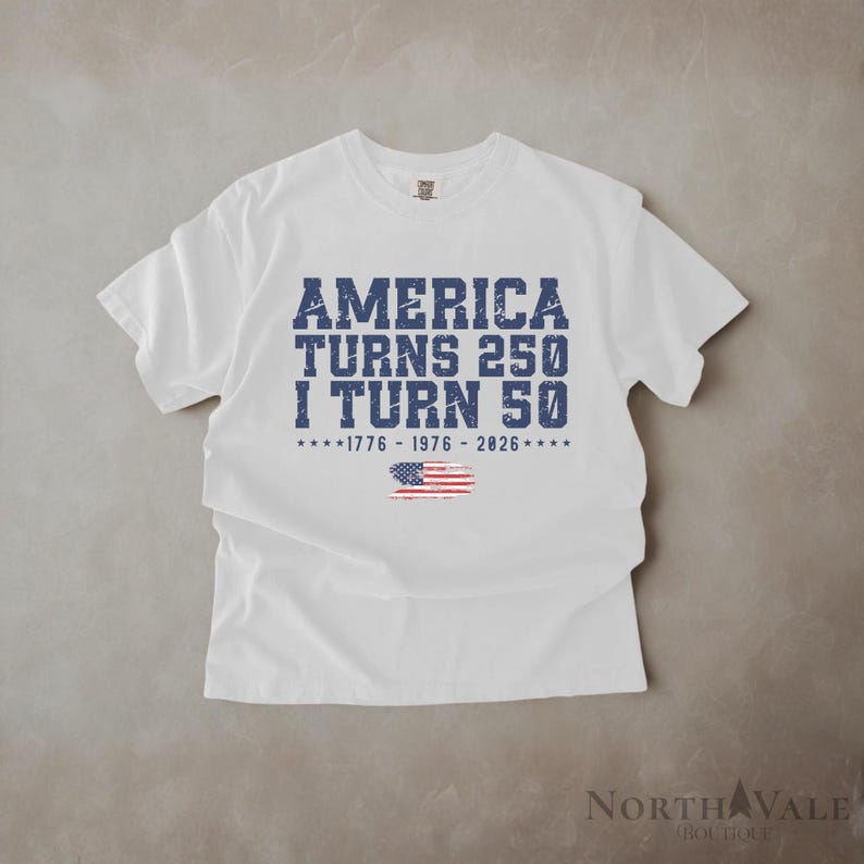 May include: White t-shirt with the text "AMERICA TURNS 250 I TURN 50" in blue, distressed font. Below the text is the American flag and the years 1776-1976-2026. The shirt is made of a soft material.