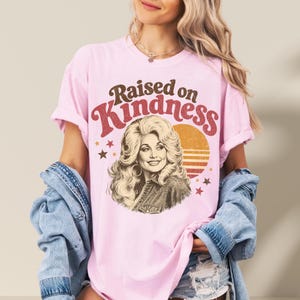 May include: A light pink short-sleeve t-shirt featuring a graphic print of a woman with voluminous blonde hair. The text "Raised on Kindness" is printed in brown and red above the portrait, accompanied by a sunset graphic and small stars.