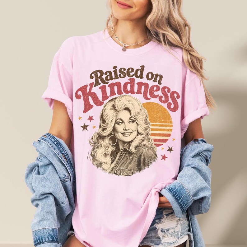 May include: A light pink short-sleeve t-shirt featuring a graphic print of a woman with voluminous blonde hair. The text "Raised on Kindness" is printed in brown and red above the portrait, accompanied by a sunset graphic and small stars.