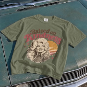 May include: An olive green t-shirt featuring a graphic print of a woman with voluminous blonde hair. The text "Raised on Kindness" is printed in a red, distressed font above the portrait, accompanied by a yellow sun graphic and small stars.