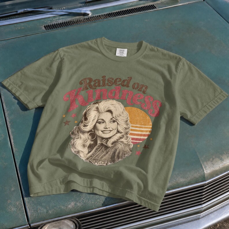 May include: An olive green t-shirt featuring a graphic print of a woman with voluminous blonde hair. The text "Raised on Kindness" is printed in a red, distressed font above the portrait, accompanied by a yellow sun graphic and small stars.