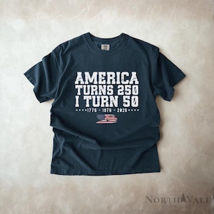 May include: Dark navy blue t-shirt with white text: "AMERICA TURNS 250 I TURN 50". Below the text is the American flag and the years 1776-1976-2026. The shirt is a crew neck and has short sleeves.