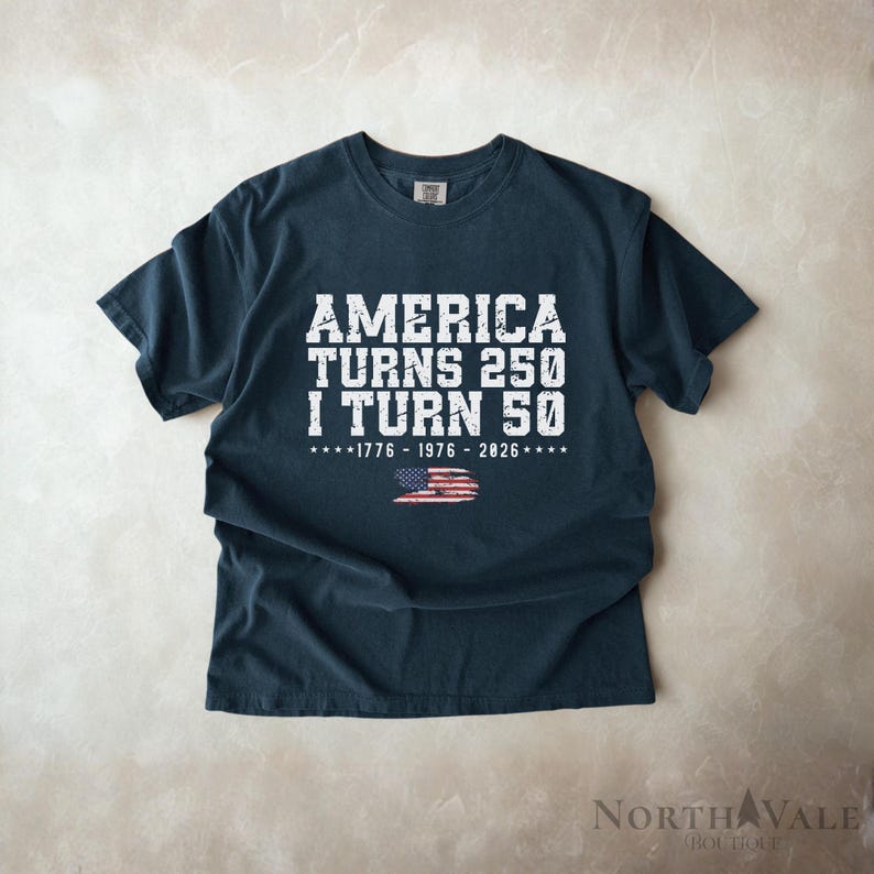 May include: Dark navy blue t-shirt with white text: "AMERICA TURNS 250 I TURN 50". Below the text is the American flag and the years 1776-1976-2026. The shirt is a crew neck and has short sleeves.