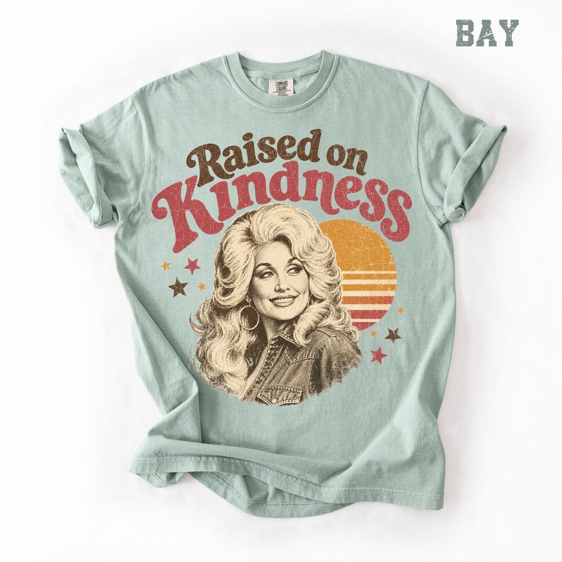 May include: A light sage green t-shirt featuring a graphic print of a woman with voluminous blonde hair. The text reads Raised on Kindness in brown and red lettering. A circular orange sunset design with horizontal stripes and small stars surrounds the portrait.