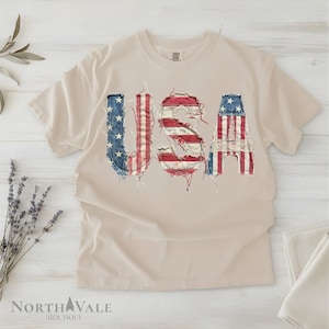May include: A beige t-shirt with the word "USA" in a distressed American flag design. The letters are made to look like they are made of denim and red and white stripes. The shirt is on a white wooden surface.