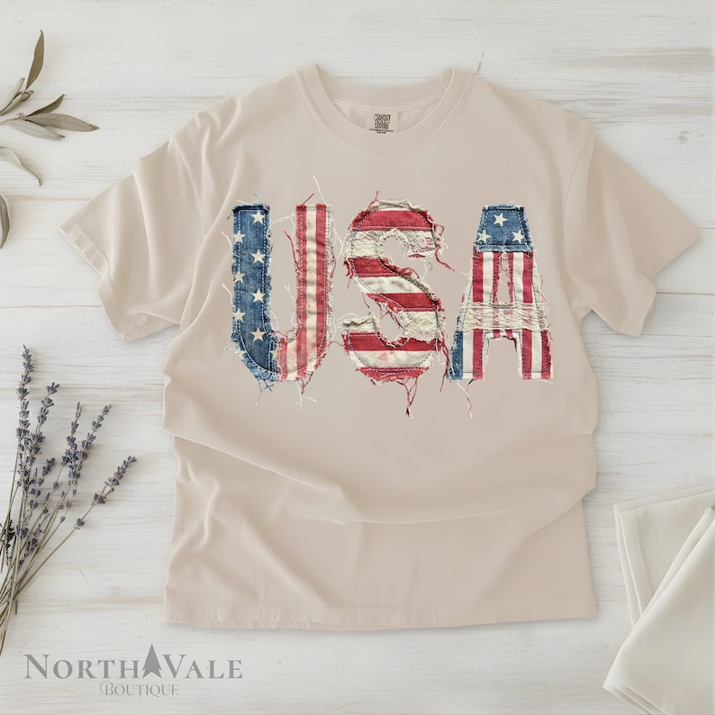 May include: A beige t-shirt with the word "USA" in a distressed American flag design. The letters are made to look like they are made of denim and red and white stripes. The shirt is on a white wooden surface.
