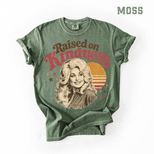 May include: A moss green short-sleeve t-shirt featuring a graphic print of a woman with voluminous blonde hair. The text "Raised on Kindness" is printed in red above the portrait, accompanied by a yellow sun graphic and small scattered stars.