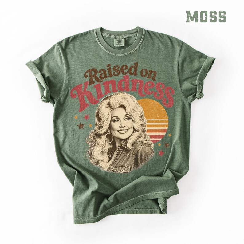May include: A moss green short-sleeve t-shirt featuring a graphic print of a woman with voluminous blonde hair. The text "Raised on Kindness" is printed in red above the portrait, accompanied by a yellow sun graphic and small scattered stars.