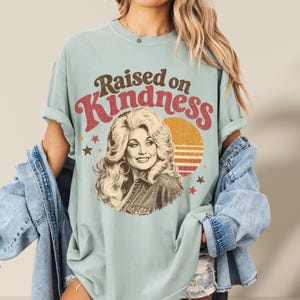 May include: A light sage green t-shirt featuring a graphic print of a woman with voluminous blonde hair. The text "Raised on Kindness" is printed in brown and red above the portrait. A yellow sun and small stars are visible in the background.