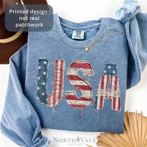 May include: A blue crewneck sweatshirt featuring a printed design of the letters USA in a distressed American flag pattern. The graphic mimics a frayed patchwork style with red, white, and blue colors. The text Printed design-not real patchwork is visible.