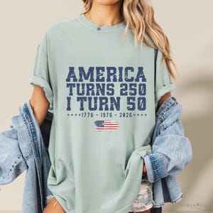 May include: A light green t-shirt with the text "AMERICA TURNS 250 I TURN 50" in dark blue. Below the text is the American flag and the years 1776-1976-2026. The shirt is a casual style.