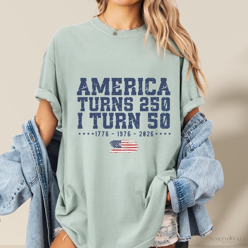 May include: A light green t-shirt with the text "AMERICA TURNS 250 I TURN 50" in dark blue. Below the text is the American flag and the years 1776-1976-2026. The shirt is a casual style.