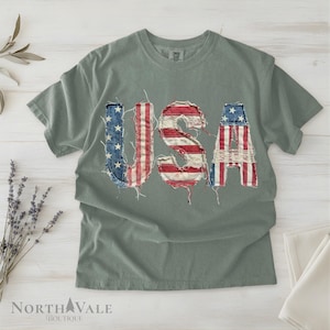 May include: A sage green t-shirt with the word "USA" appliqued across the front. The letters are made from distressed denim and fabric in the colors of the American flag. The shirt is a casual, patriotic style.