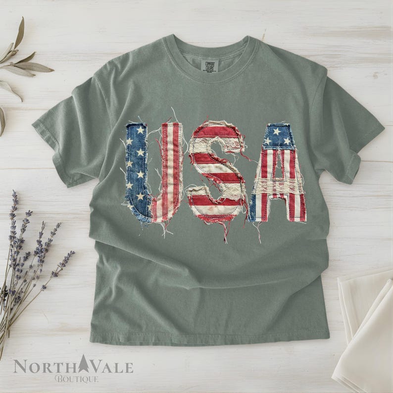 May include: A sage green t-shirt with the word "USA" appliqued across the front. The letters are made from distressed denim and fabric in the colors of the American flag. The shirt is a casual, patriotic style.
