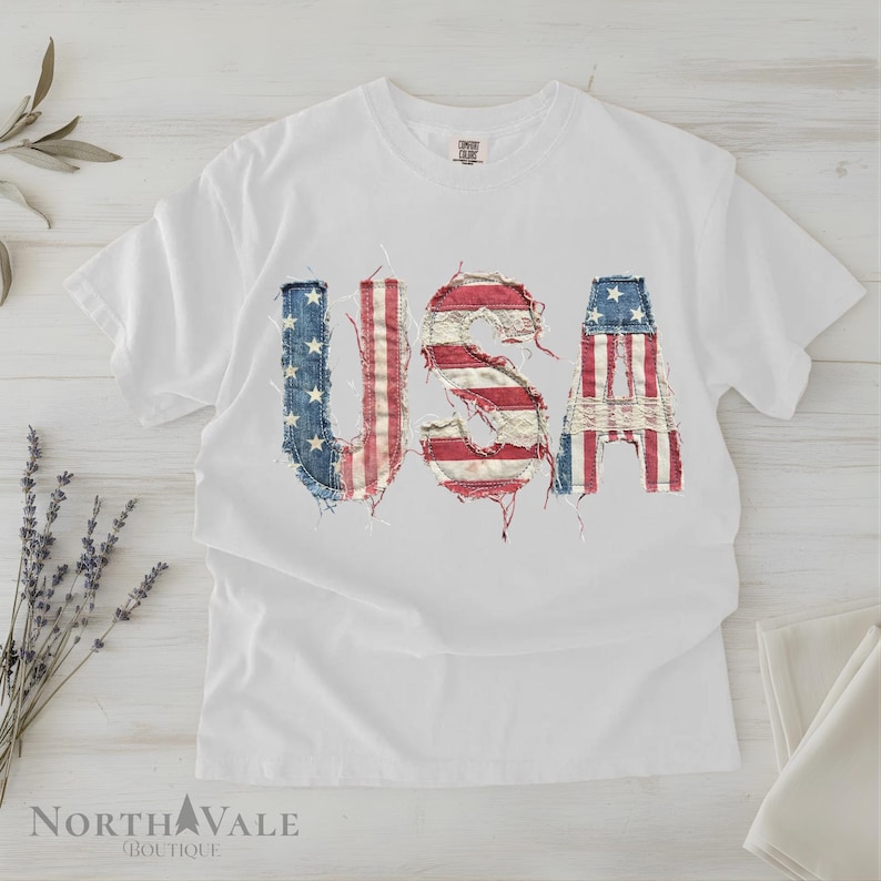 May include: White t-shirt with the word "USA" in distressed, patriotic flag-themed lettering. The letters are made from patches of red, white, and blue fabric, giving a vintage look. The shirt is laid flat on a light-colored surface.