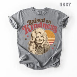May include: A grey short-sleeve t-shirt featuring a graphic print of a woman with voluminous blonde hair. The text "Raised on Kindness" is arched above the portrait in brown and red. A sunset graphic and small stars surround the central figure.