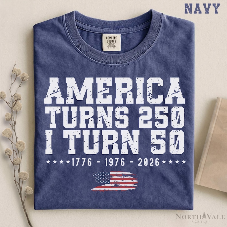May include: Navy blue t-shirt with white text: "AMERICA TURNS 250 I TURN 50" and the years 1776, 1976, and 2026. A distressed American flag graphic is at the bottom. The shirt is from North Vale Boutique.
