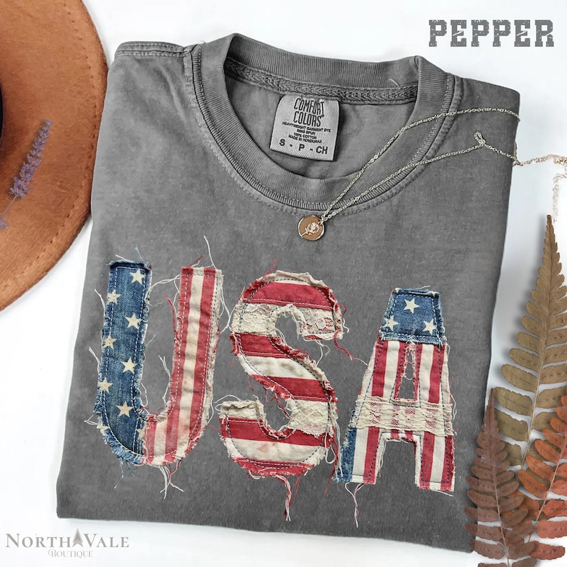 May include: Gray Comfort Colors t-shirt with a distressed "USA" design in red, white, and blue. The letters are made from patches of denim and fabric, with a frayed edge. A silver necklace with a pendant is draped across the shirt.