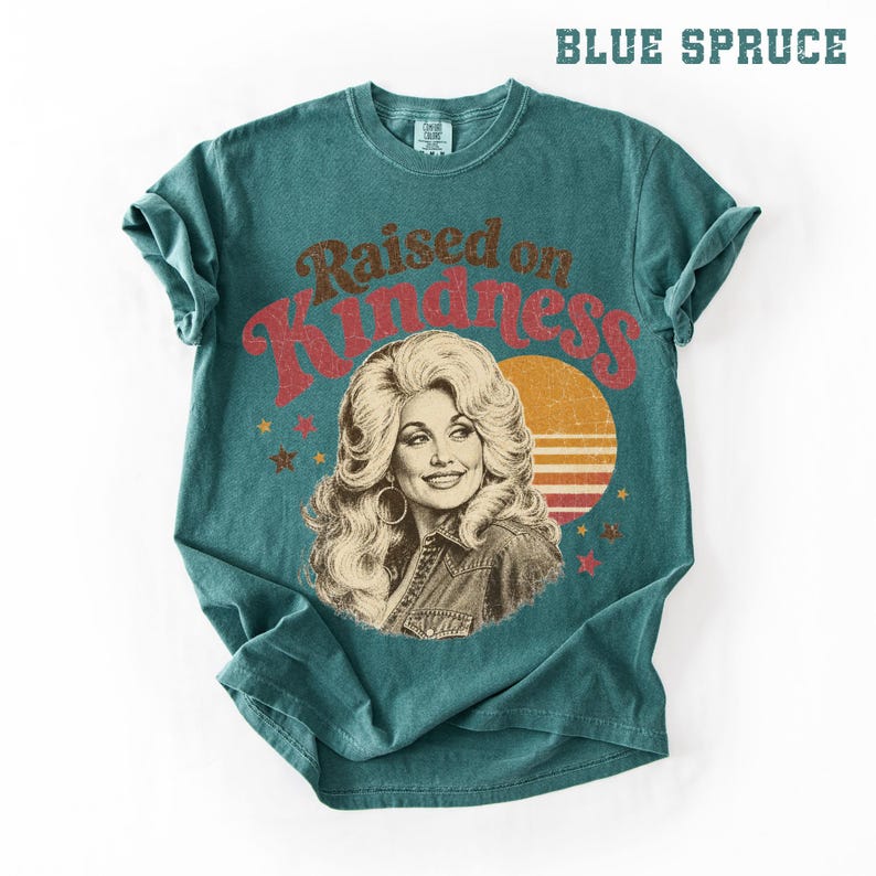 May include: A blue spruce colored short-sleeve t-shirt featuring a graphic print of a woman with voluminous blonde hair. The text "Raised on Kindness" is printed in a distressed red font above the portrait, accompanied by a sunset and stars.