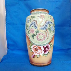 May include: A ceramic vase featuring a raised relief design of a mythical bird with blue wings and a long tail, surrounded by green vines and small flowers. Below the bird, large pink and deep red roses are painted on a gradient tan background.