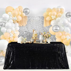 May include: A black sequin tablecloth covers a rectangular table, set for a celebration. The backdrop features a mosaic wall, balloons in white, silver, and peach, and decorative elements like cakes and flowers. The scene suggests a party or event setting.