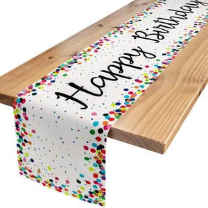 May include: White birthday table runner with colorful confetti and the words "Happy Birthday" in black script. The runner is draped over a wooden table, creating a festive look for a birthday celebration.