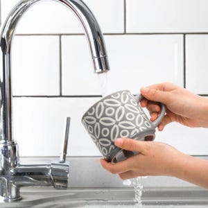 May include: A gray ceramic mug with a white floral pattern is held under a stream of water from a chrome faucet. The mug has a handle and is being washed in a white-tiled sink.