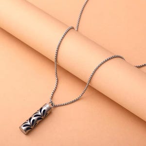 May include: A silver-colored necklace with a cylindrical pendant. The pendant features a dark blue interior and a decorative pattern of stylized leaves. The chain is a fine, linked design. The background is a light peach color.