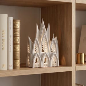 Pode incluir: Uma escultura decorativa de cerâmica branca em forma de catedral gótica com arcos pontiagudos e detalhes recortados intrincados. A estrutura é iluminada por dentro, projetando um brilho suave através das aberturas arquitetônicas na prateleira.