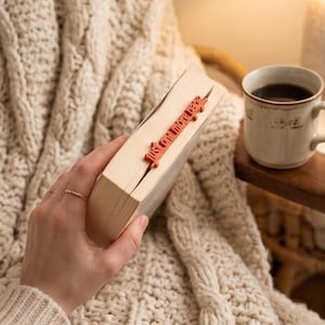 Op de afbeelding: Een boek met een terracotta-kleurige boekenlegger die uit de bovenrand steekt, voorzien van de lasergesneden tekst "just one more page" in een cursief lettertype. Het boek heeft crèmekleurige pagina's en wordt vastgehouden door een hand met een dunne gouden ring.