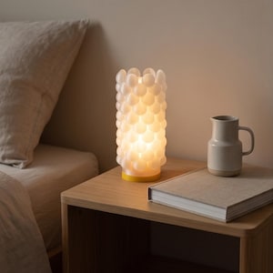 Könnte beinhalten: Eine zylindrische Tischleuchte mit einer durchscheinenden weißen Oberfläche, die aus zahlreichen kleinen, abgerundeten, blasenartigen Vorsprüngen besteht. Die Lampe steht auf einem gelben Sockel und strahlt warmes Licht aus.