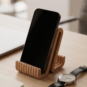 Op de afbeelding: Een zwarte smartphone rustend op een lichtbruine, U-vormige telefoonstandaard met een geribbelde, getextureerde basis. De standaard heeft een gebogen, verticale steunstructuur die het apparaat rechtop in een hoek houdt voor stabiliteit.