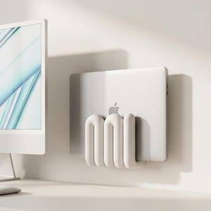 Può includere: Un supporto da parete per laptop di colore bianco con un design tubolare arrotondato. Il supporto è fissato a una parete e sostiene un laptop argentato con logo Apple. La struttura è composta da tre anelli verticali e curvi.