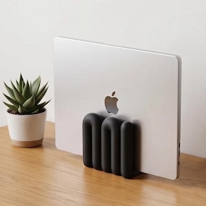 Può includere: Un computer portatile argentato con logo Apple è tenuto in posizione verticale da un supporto nero opaco a forma di U con diverse nervature. Accanto al portatile si trova una piccola pianta succulenta verde in un vaso bianco e beige.