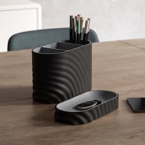 Pode incluir: Um organizador de secretária preto com uma textura exterior ondulada e vários compartimentos interiores contendo várias canetas, emparelhado com um tabuleiro oval preto a condizer de perfil baixo contendo um pequeno dispositivo eletrónico.