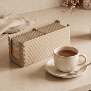 Puede incluir: Un organizador rectangular de color beige para bolsitas de té con un patrón geométrico texturizado, que contiene varias bolsitas etiquetadas como Artisan Tea Pure Chamomile. Al lado hay una taza de té de cerámica beige a juego sobre un plato.