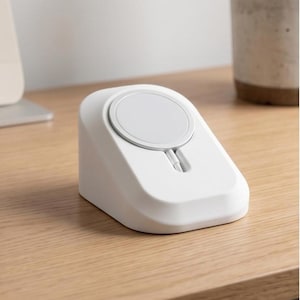 Puede incluir: Una base de carga inalámbrica blanca en forma de cuña con una almohadilla de carga magnética circular integrada en la superficie frontal inclinada. El dispositivo tiene un acabado mate liso e incluye una pequeña ranura para el cable.