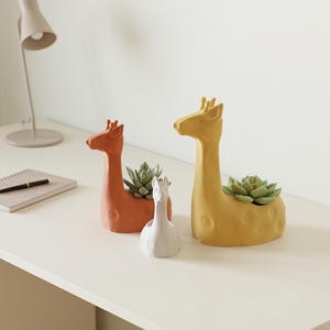 May include: Three giraffe-shaped planters in orange, white, and yellow, each holding a succulent plant. The planters are made of ceramic and have a smooth finish. The giraffe planters are of different sizes and are placed on a white surface.