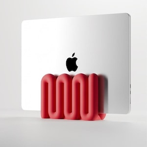 Puede incluir: Un portátil plateado con un logotipo de Apple negro se mantiene en posición vertical mediante un soporte rojo mate con forma de onda serpenteante. El soporte presenta un diseño continuo de curvas en S que proporciona una base estable.