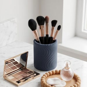 May include: A collection of makeup brushes with black bristles and rose gold handles in a navy blue wavy-patterned holder. A gold eyeshadow palette, a decorative tray with cotton pads, and a perfume bottle are also on the marble surface.