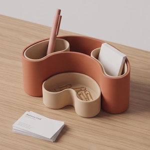 Pode incluir: Um organizador de secretária em terracota e bege com compartimentos curvos contendo uma caneta rosa, um bloco de notas autocolantes brancas e clipes dourados. Uma pequena pilha de cartões de visita brancos com o texto Business Card à frente.